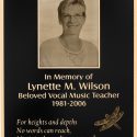 Memorial plaque featuring a portrait of Lynette M. Wilson with the inscription 'In Memory of Lynette M. Wilson, Beloved Vocal Music Teacher 1981-2006' and a quote 'For heights and depths, No words can reach.' accompanied by an engraved dragonfly design on a dark background."