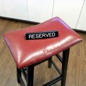 Red cushioned stool with a custom metal sign reading 'RESERVED' placed on top, set against a wooden floor and white cabinetry background.